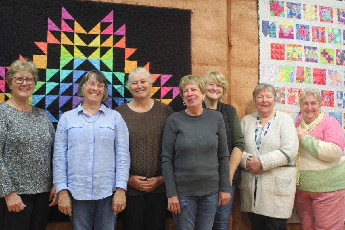Members of the Gilgandra Wednesday Patchworkers and Quilters stand together in front of two colourful quilts.